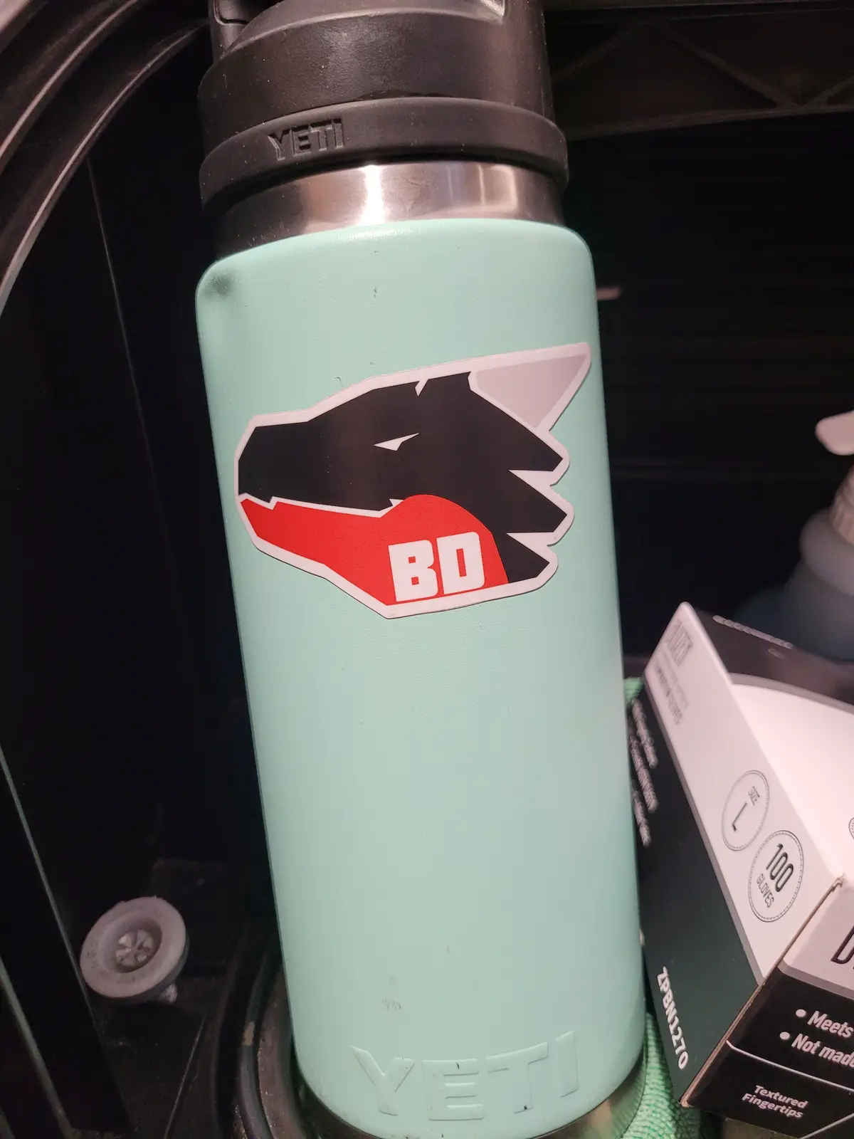 A light green Yeti water bottle with a black and red sticker of a stylized wolf head, with the letters 'BD' in white inside…