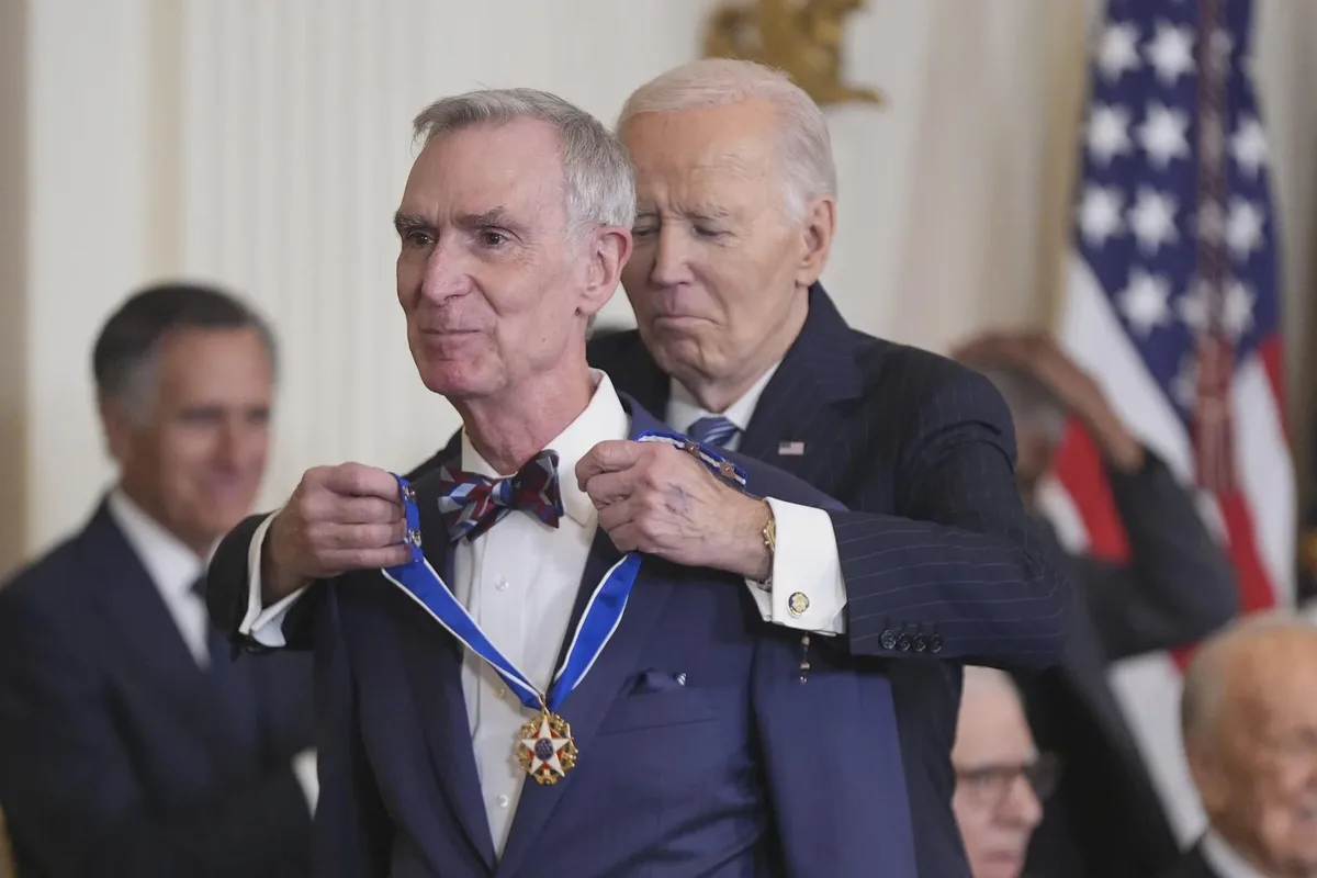 A man in a blue suit and patriotic bow tie is being awarded a medal by President Joe Biden, who is visibly frowning and…