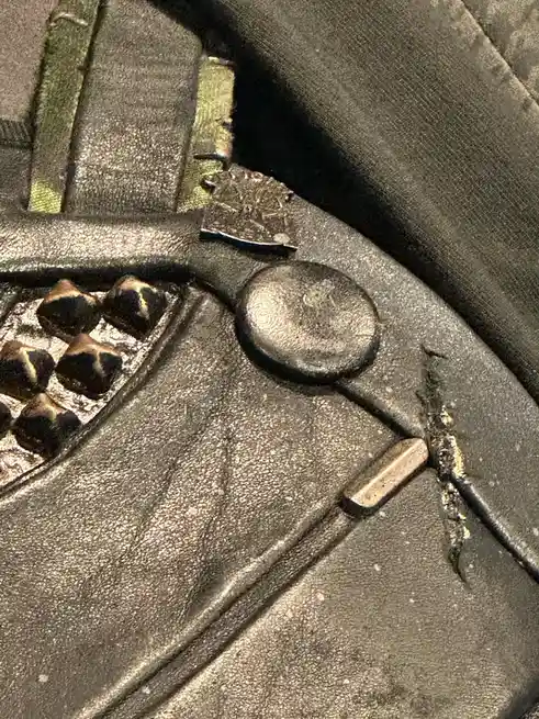 A close-up photo of a worn, textured black leather bag or jacket with metal hardware, including a circular button, rivets…
