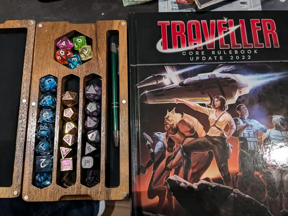 A wooden dice tray filled with colorful polyhedral dice sits next to the 'Traveller Core Rulebook Update 2022' book, which…