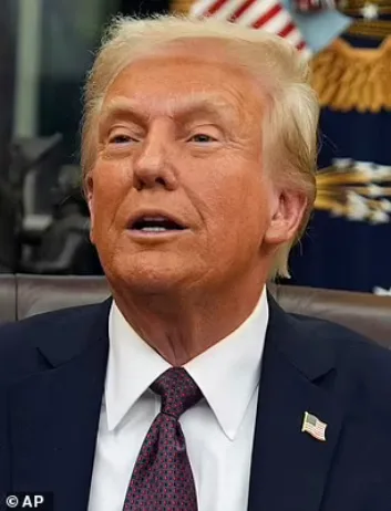 a close-up photo of Donald Trump in a dark suit and red patterned tie, sitting in an office with an american flag pin on his…
