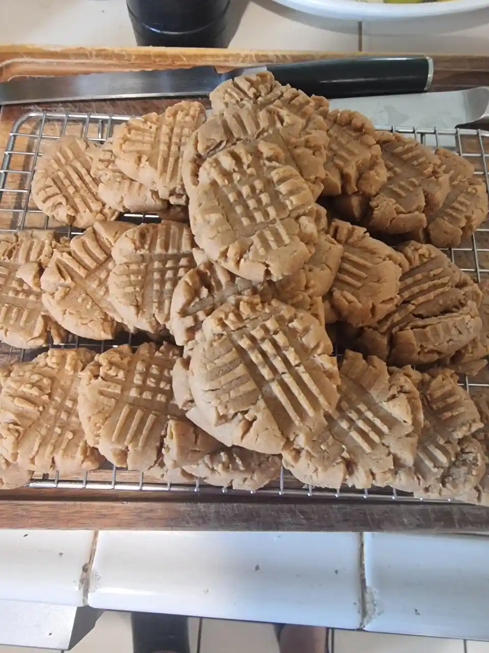 A close-up photo shows a large pile of freshly baked peanut butter cookies with the classic criss-cross pattern, resting on…