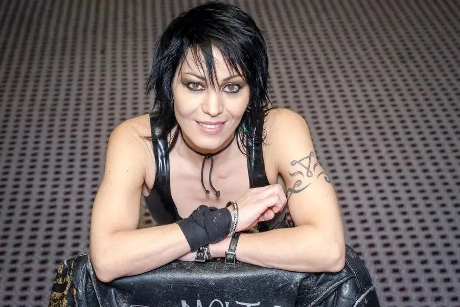 A woman with short black hair, dark eye makeup, and a black leather top leans forward with her arms crossed on a black…