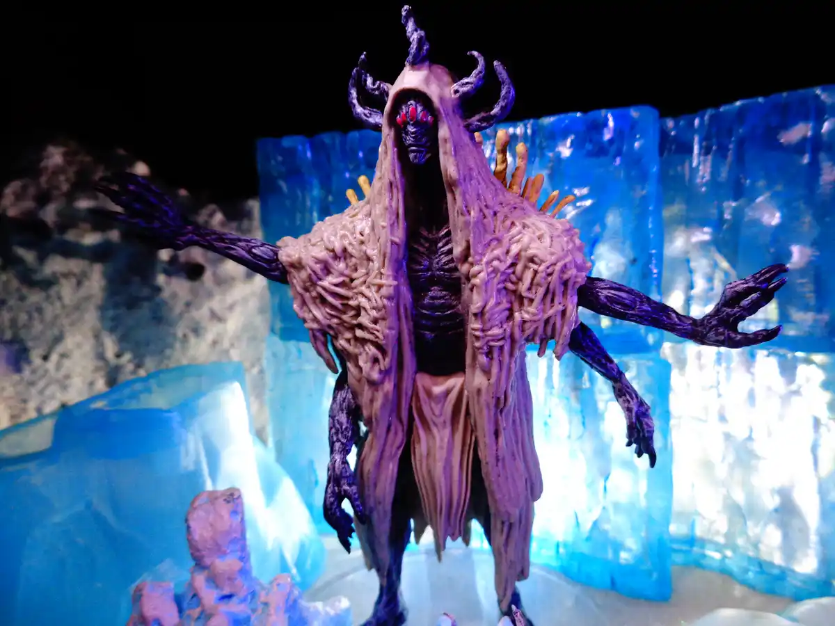 A grotesque, purple-hued monster figurine with horns, a hooded, noodle-like robe, and multiple limbs stands with arms…