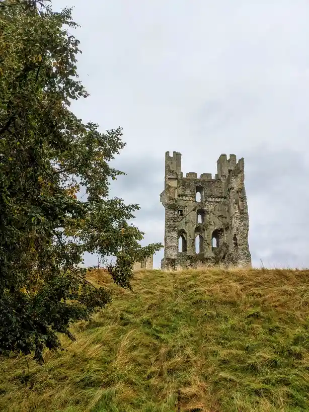A photograph shows the weathered, stone ruins of a castle tower standing atop a grassy hill under an overcast sky, with tree…