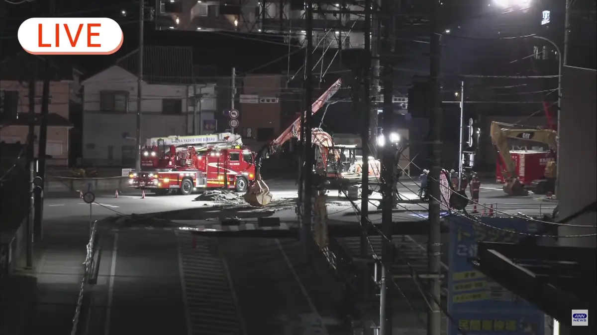 A nighttime live news broadcast image shows emergency vehicles, including a red fire truck and excavators, at a street scene…