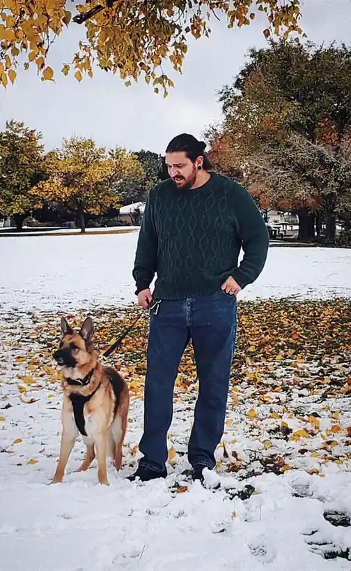 A man in a green sweater and jeans stands in a snowy park with autumn leaves, holding a leash attached to a German Shepherd…