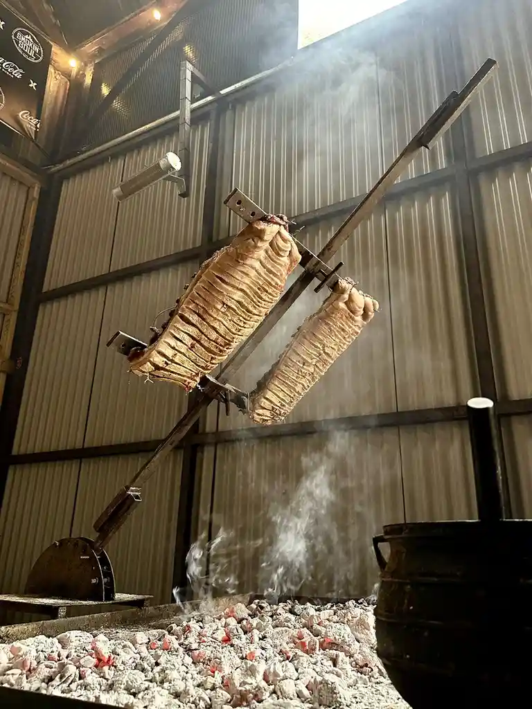 Two large, roasted pork carcasses hang on a spit over glowing embers in a rustic, smoke-filled kitchen. The meat is visibly…