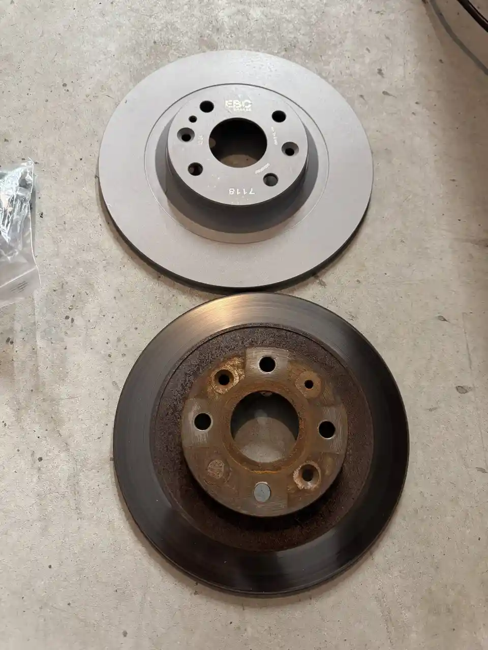 Two brake rotors lie on a concrete floor, one shiny new EBC Brakes rotor above and one rusty, worn-out rotor below. The…