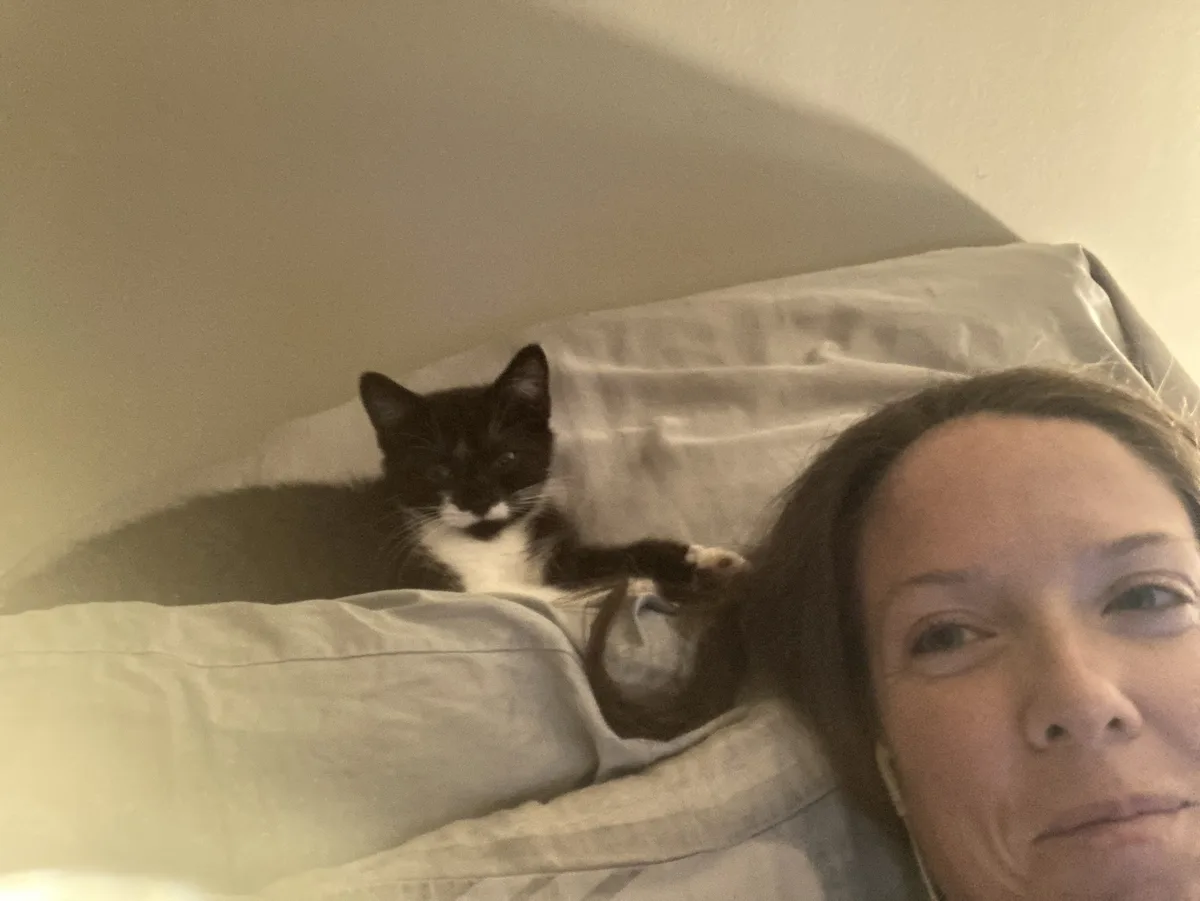 A woman with brown hair lies in bed, looking at the camera with a tired expression, while a black-and-white cat rests on a…