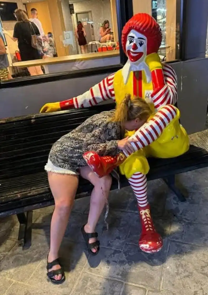 A woman is visibly crying while sitting on a bench next to a large Ronald McDonald statue, her head resting on his arm. She…