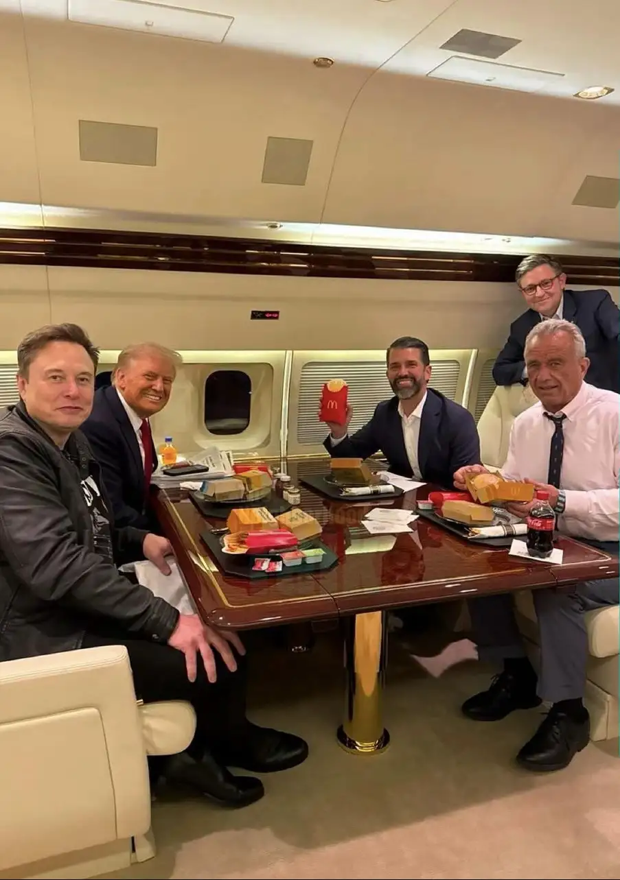 Four men, including Elon Musk and Donald Trump, are seated around a polished wooden table inside a luxurious private jet…