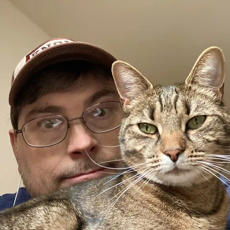 A man wearing glasses and a baseball cap holds a tabby cat close to his face in a selfie. The cat has green eyes and a calm…