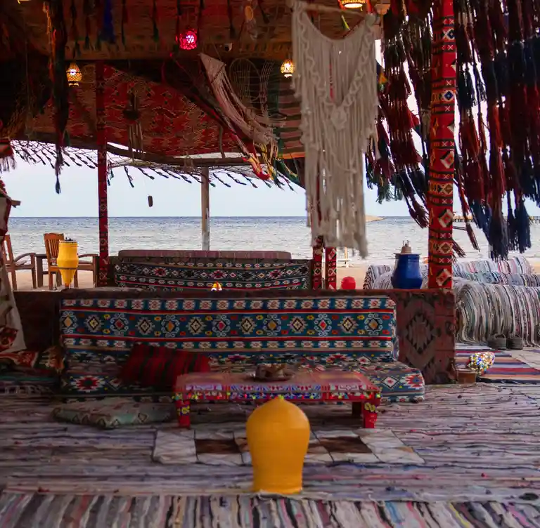 A vibrant, bohemian-style beachside pavilion decorated with colorful patterned textiles, macrame, and hanging tassels…