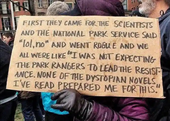 A person in a purple jacket and black glove holds a handmade cardboard sign at an outdoor protest. The sign features…