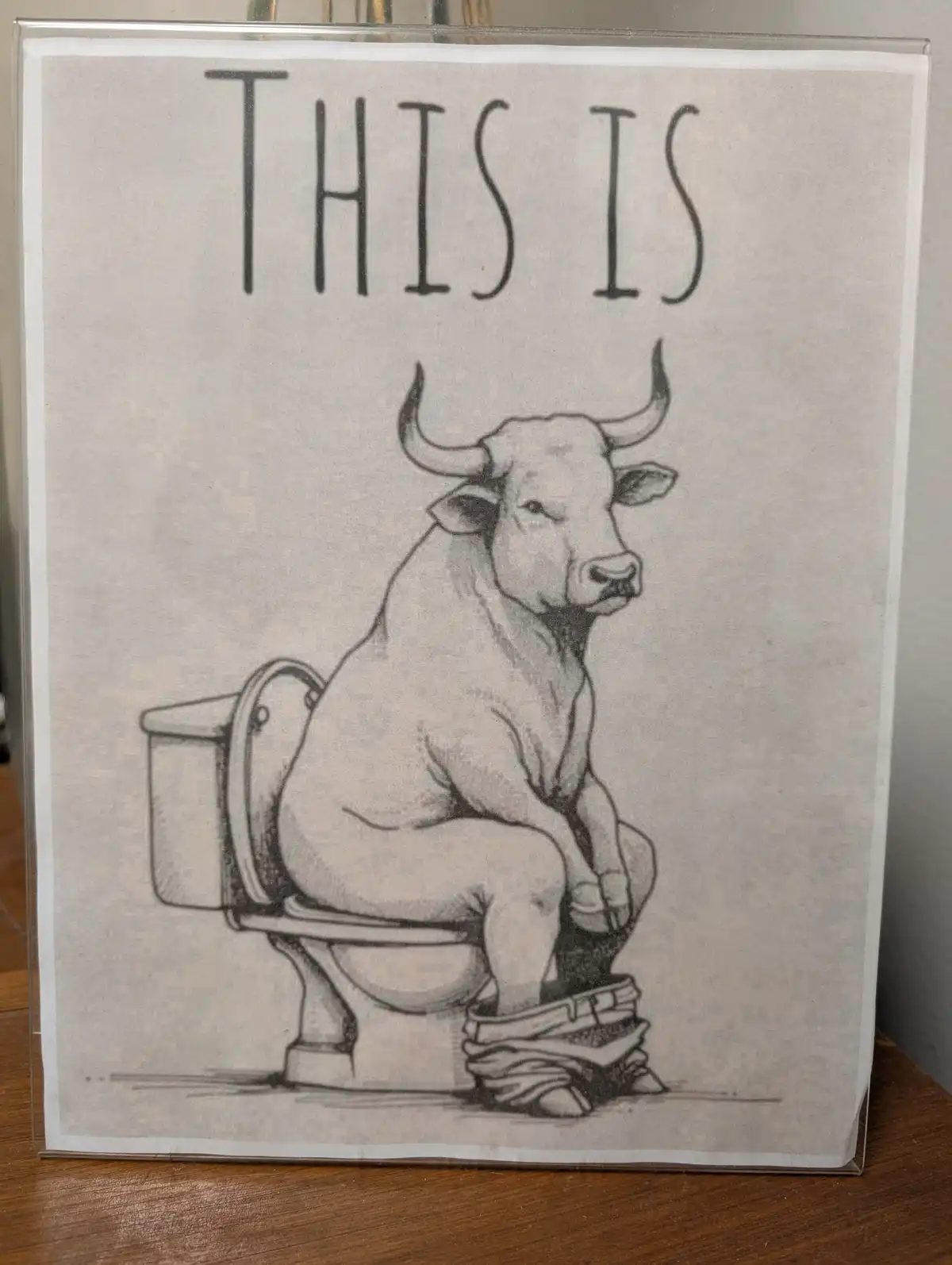 A black-and-white pencil drawing shows a bull sitting on a toilet, wearing pants and shoes, with the text 'THIS IS' above…