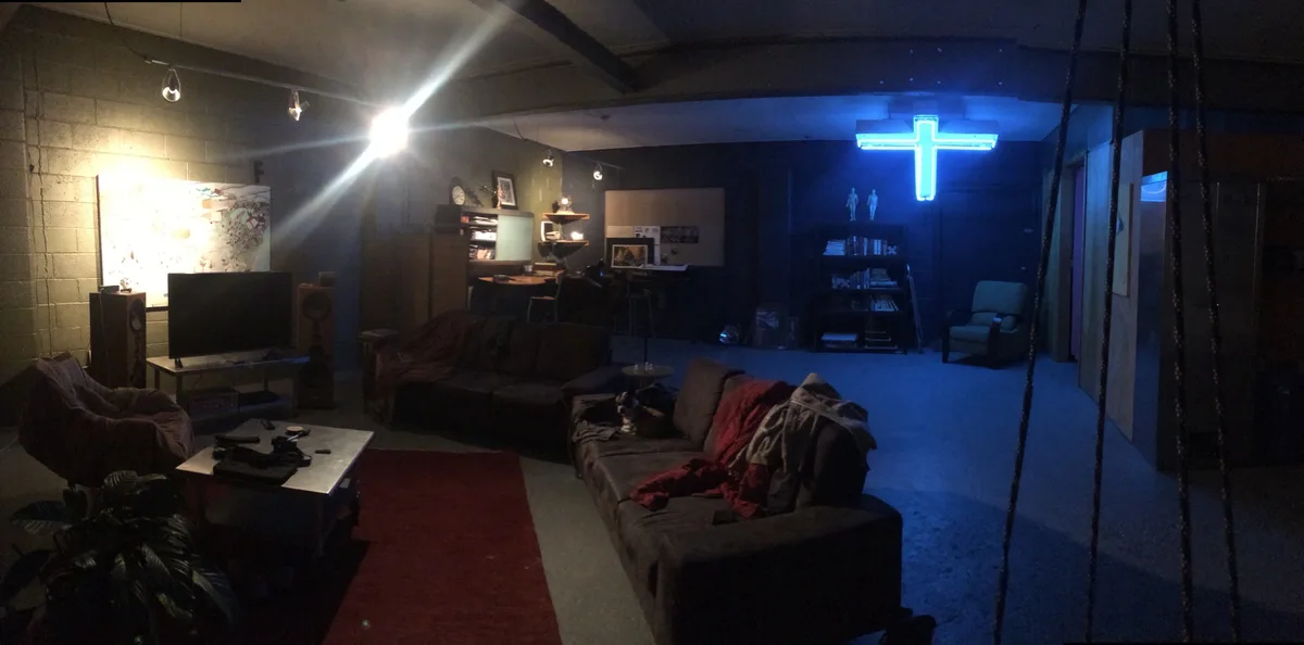 A dimly lit, cluttered basement living room features a bright blue neon cross hanging on the far wall, contrasting with warm…