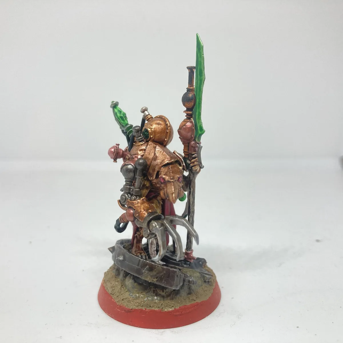 A heavily painted miniature figurine of a fantasy warrior with copper-colored armor, red eyes, and green blades, standing on…