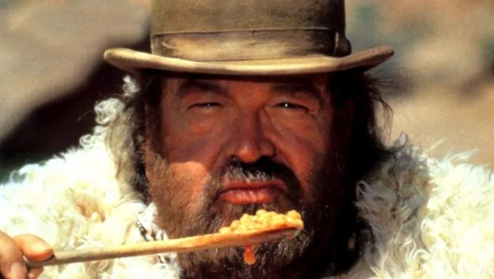 a close-up of a man with a beard and a wide-brimmed hat, wearing a fluffy white coat, holding a wooden spoon with beans on…