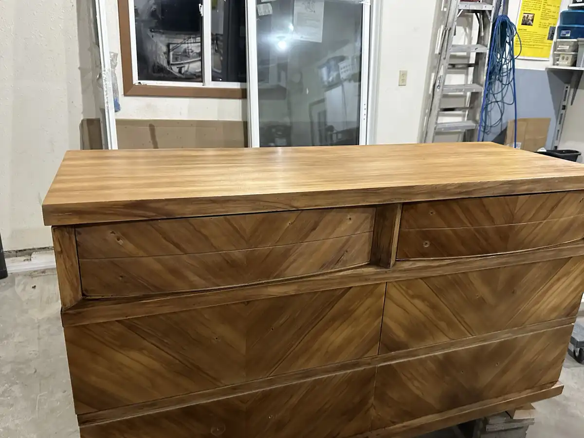 A large wooden dresser with a smooth, light-brown finish and distinctive chevron-patterned drawers is photographed in what…