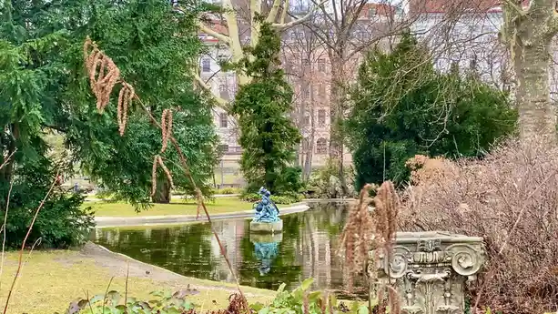 A serene park scene with a pond, a blue statue in the center, and ornate stone fixtures, framed by trees and dried plant…
