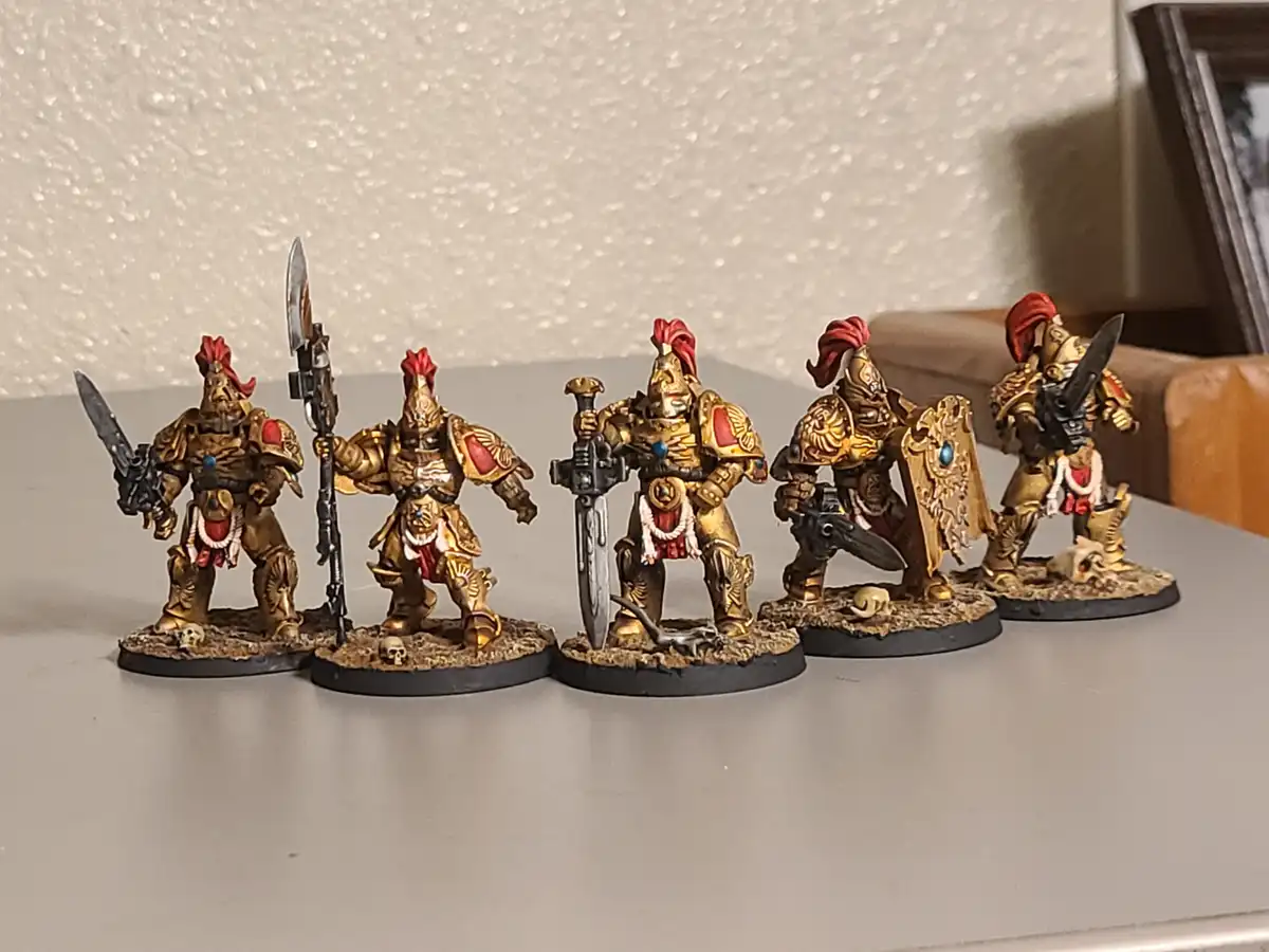 Five intricately painted miniature fantasy warriors in golden armor with red plumes stand in a row on a tabletop. Each…