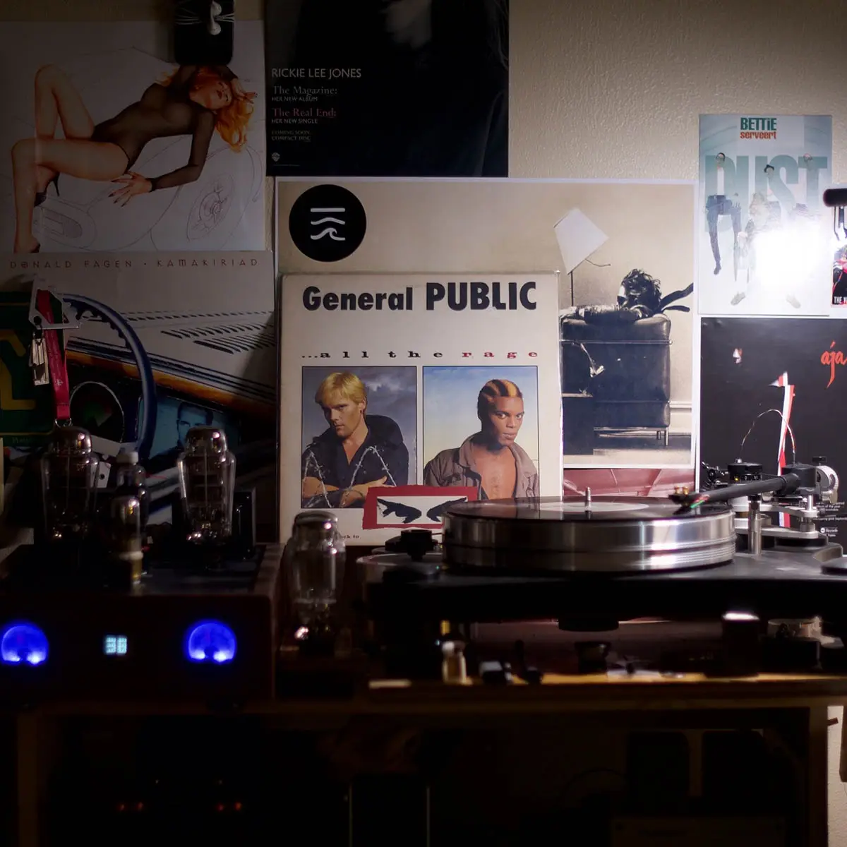 A dimly lit room features a turntable with a vinyl record on it, surrounded by various music posters including 'General…