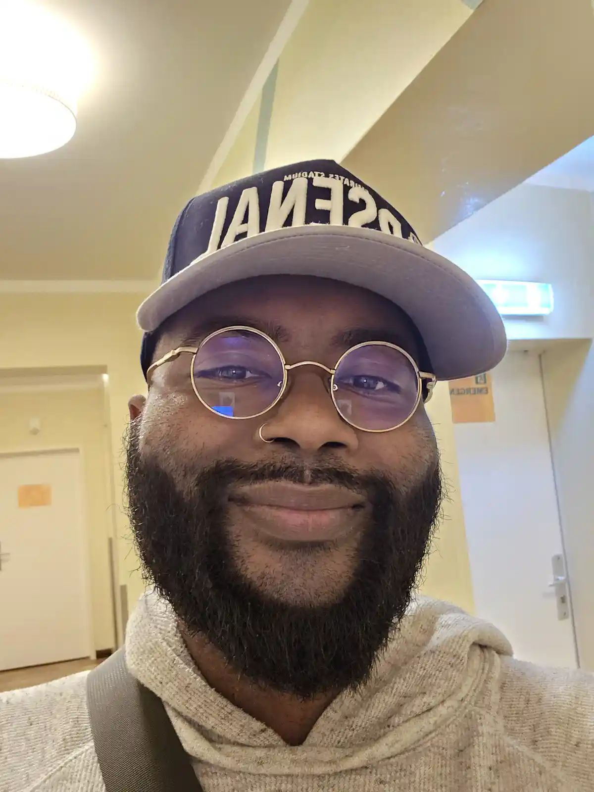 A man with a beard, round glasses, and a backwards cap smiles at the camera in a hallway. The cap reads 'OSSENAI' and he…