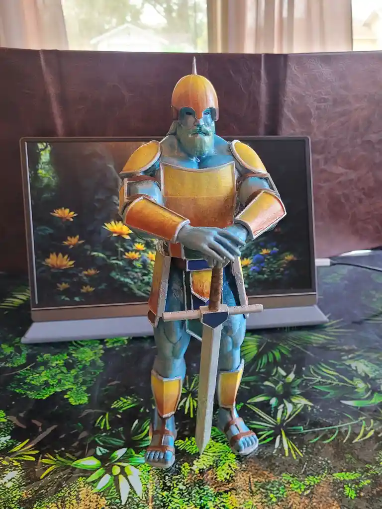 A hand-painted blue-skinned knight figurine in orange and blue armor stands holding a sword, positioned in front of a laptop…