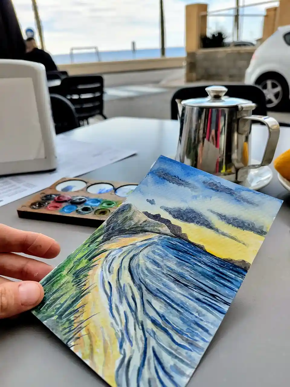 A hand holds up a watercolor painting of a coastal scene with waves and cliffs, placed on a cafe table next to a paint…