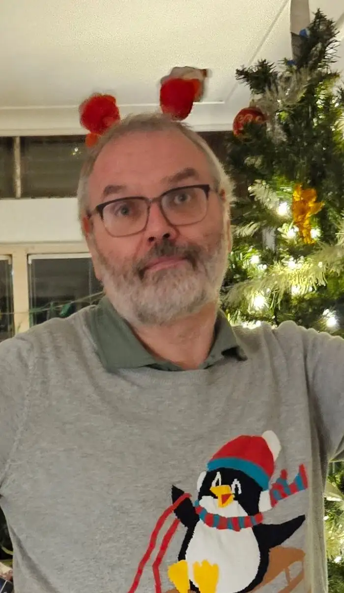 A middle-aged man with glasses and a grey sweater featuring a penguin wearing a hat and scarf looks at the camera with a…
