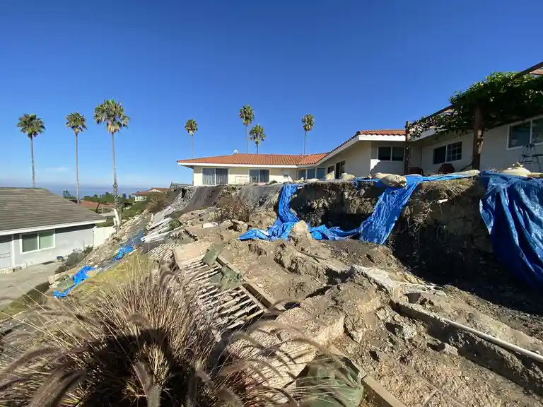 A house sits atop a severely eroded hillside, with large sections of dirt and debris having collapsed, revealing exposed…