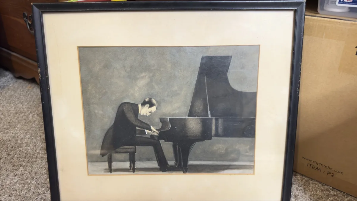 A framed black-and-white painting depicts a man in a suit leaning over a grand piano, seemingly playing or adjusting it. The…