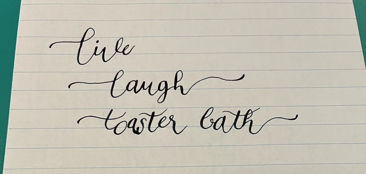Handwritten cursive text on lined notebook paper reads 'live', 'laugh', and 'toaster bath', each word on a separate line…