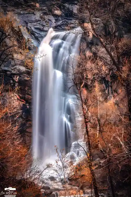 A long-exposure photograph of a waterfall cascading down a rocky cliff, framed by bare trees with autumnal orange and brown…