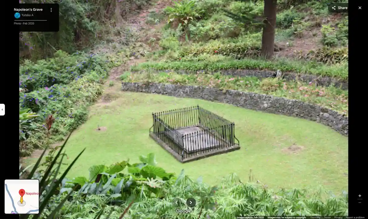 A screenshot of a Google Maps Street View image showing a simple, fenced grave in a grassy, overgrown garden with stone…