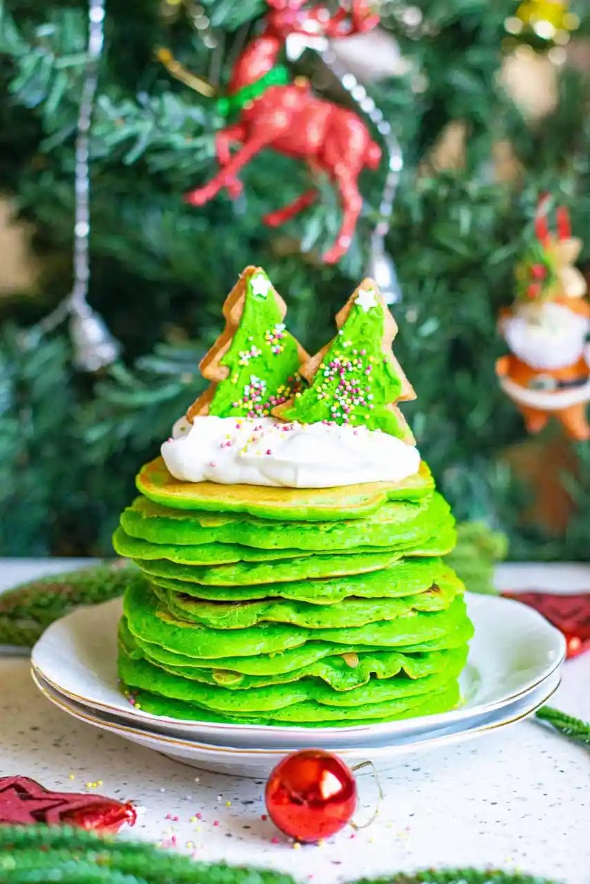 A tall stack of bright green pancakes is arranged on a white plate to resemble a Christmas tree, topped with white whipped…