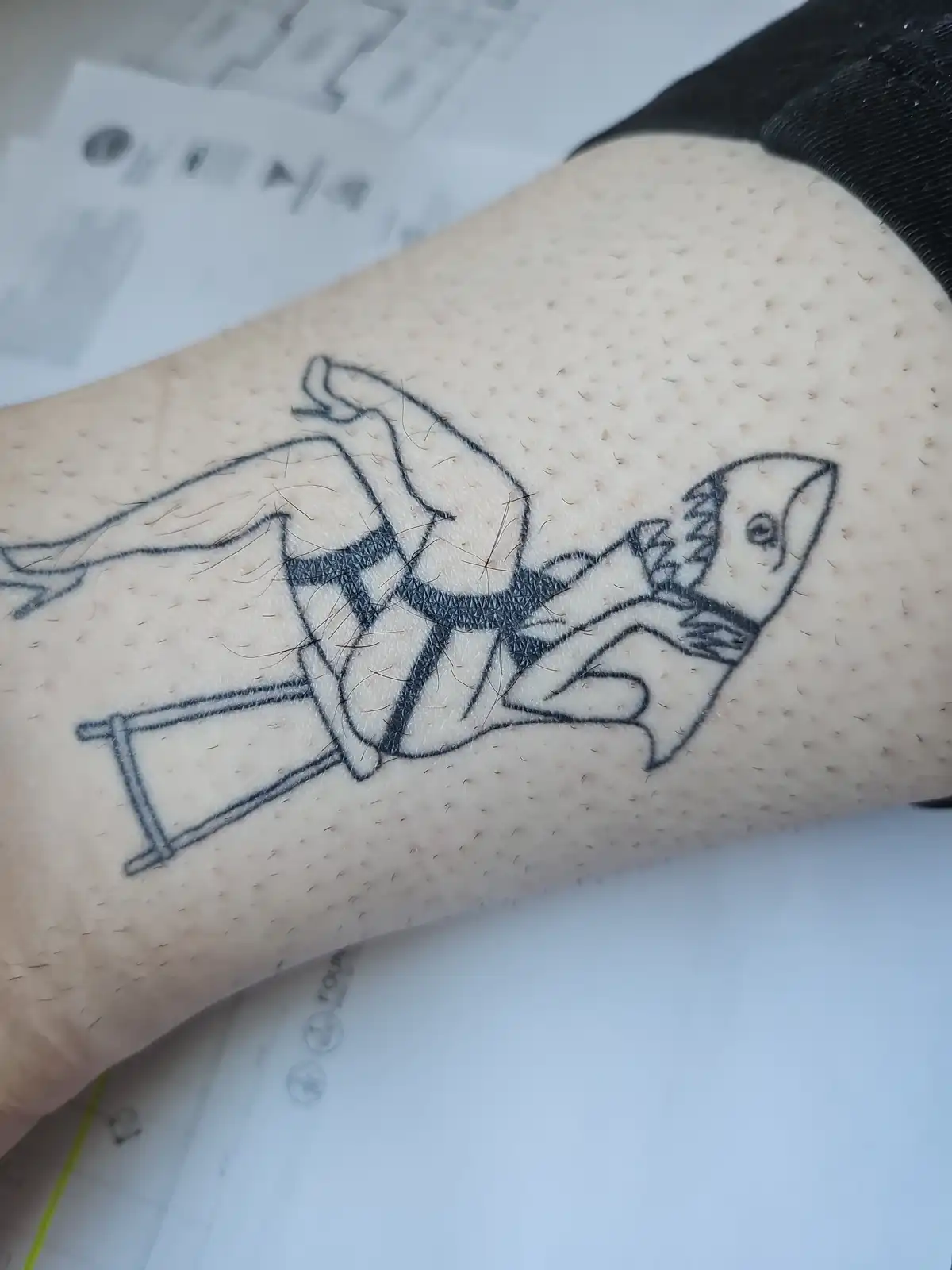 A close-up photo shows a freshly inked black-and-white tattoo on a person's forearm, depicting a cartoonish shark wearing a…