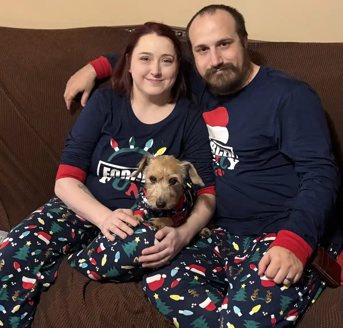 A man and woman in matching Christmas-themed pajamas sit on a brown couch with a small dog on the woman's lap. The dog wears…