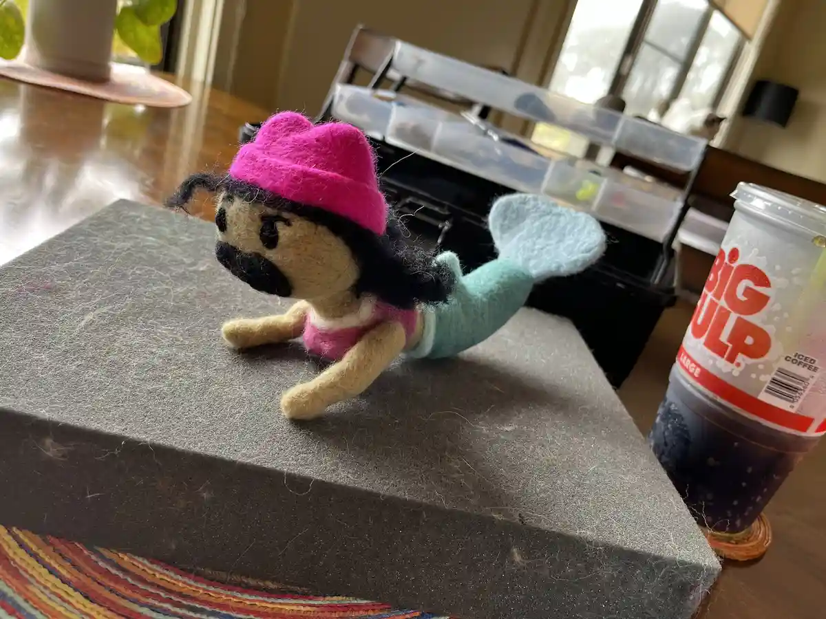 A handmade felt pug doll with a mermaid tail, wearing a pink hat and top, is posed on a gray foam block next to a large 'Big…