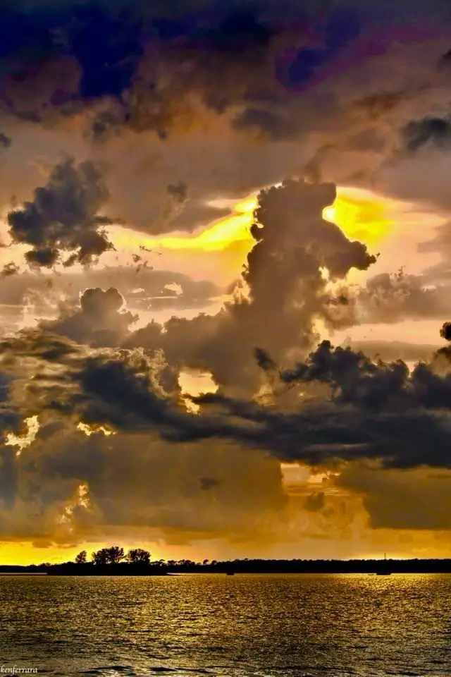 A dramatic sunset over water with dark, textured clouds partially obscuring a bright yellow sun. The horizon shows…