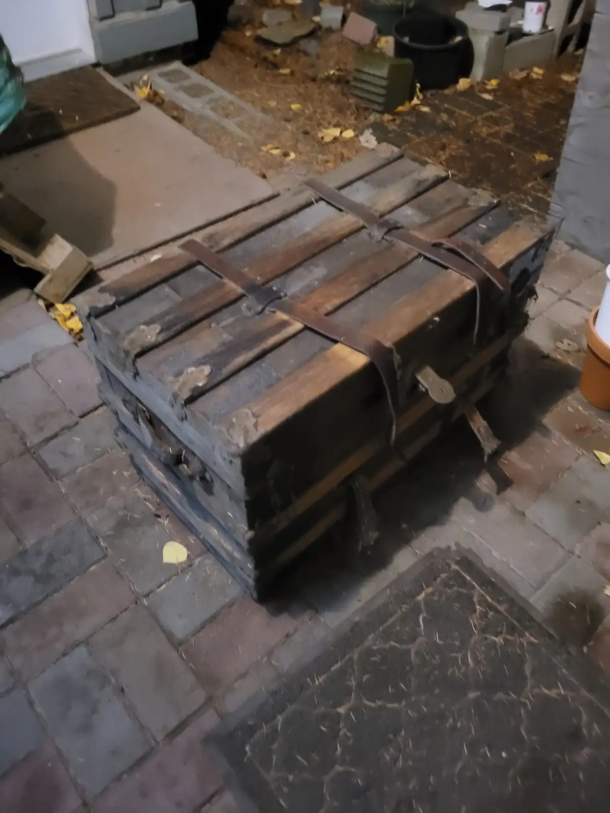 An old, weathered wooden chest with leather straps and metal latches sits on a paved patio surrounded by fallen leaves. The…
