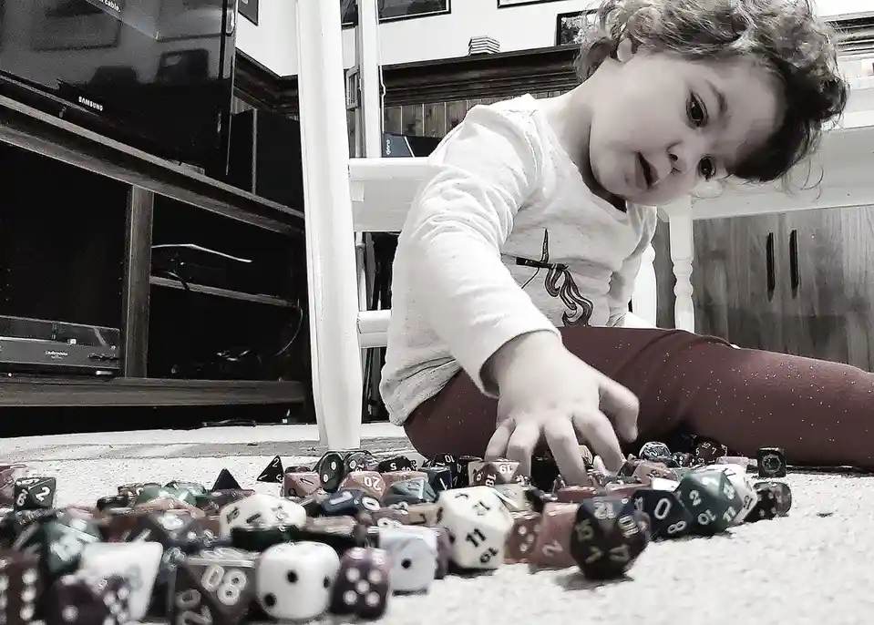 A young child sits on a carpeted floor surrounded by a large pile of colorful polyhedral dice, reaching toward them with a…