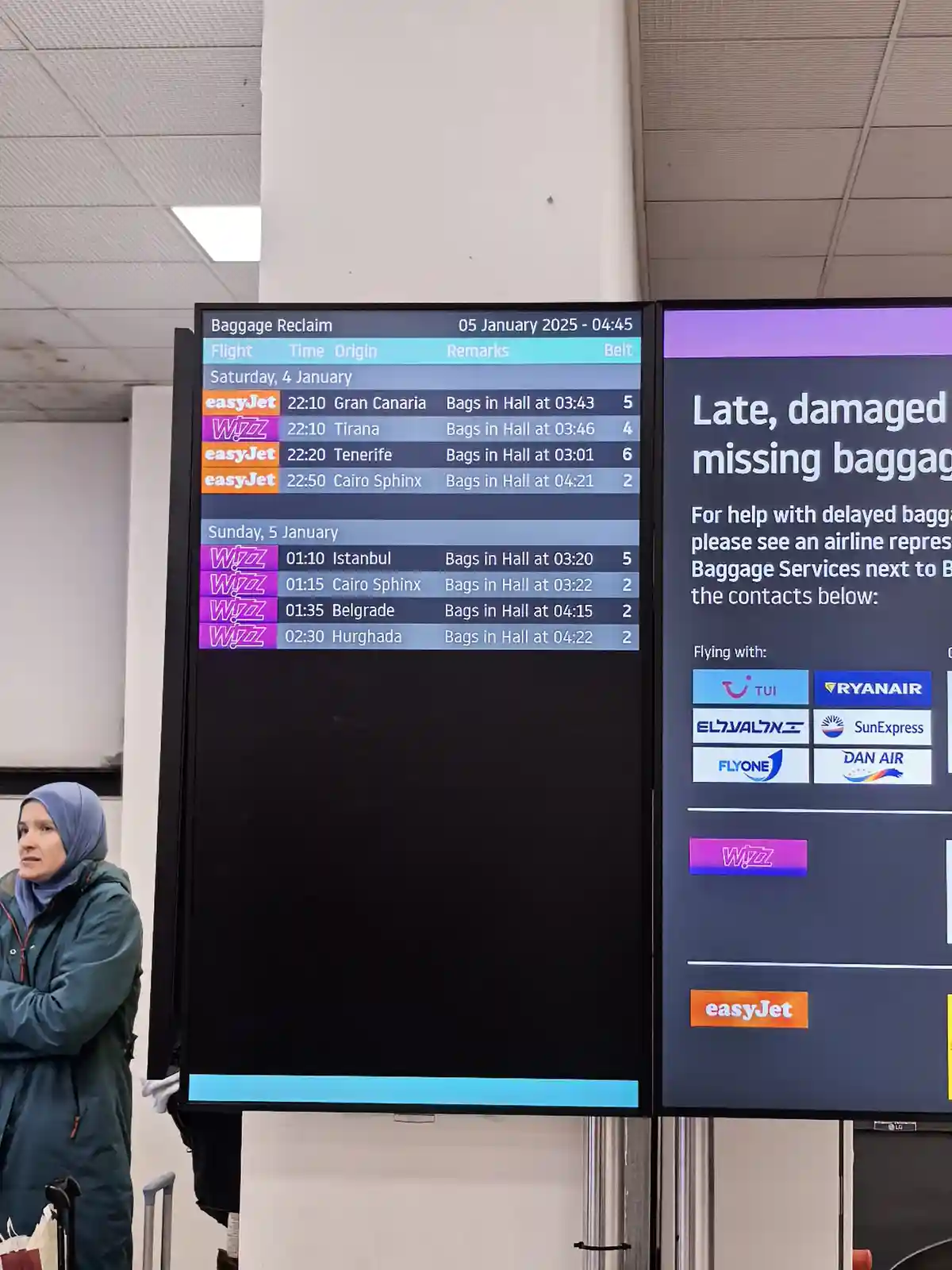 An airport baggage reclaim screen shows flights from Wizz Air and easyJet with 'Bags in Hall at' times, while a woman in a…
