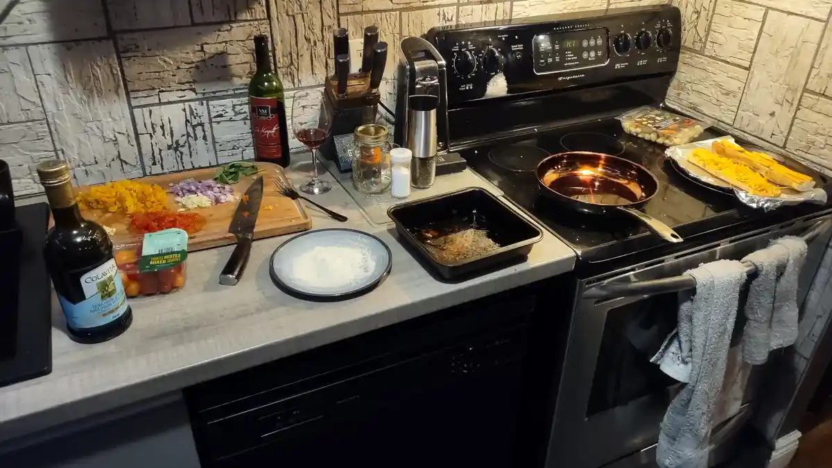 A cluttered kitchen counter and stove show ingredients like chopped onions, tomatoes, cheese, olive oil, and wine, alongside…
