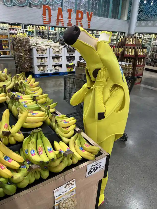 A person in a full-body yellow banana costume stands in a grocery store aisle next to a banana display, hands covering their…