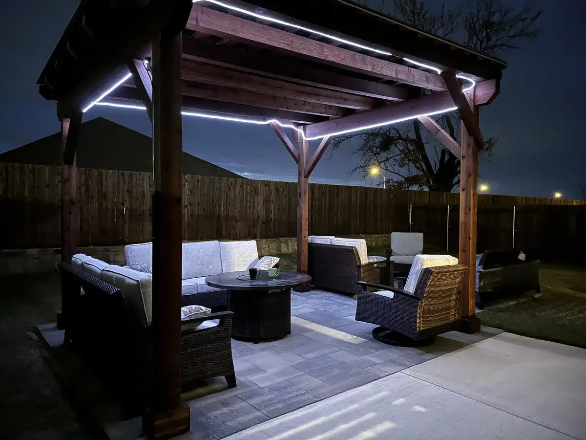 A nighttime photo of a well-lit backyard patio with a wooden pergola, wicker furniture, and a stone patio. The pergola beams…