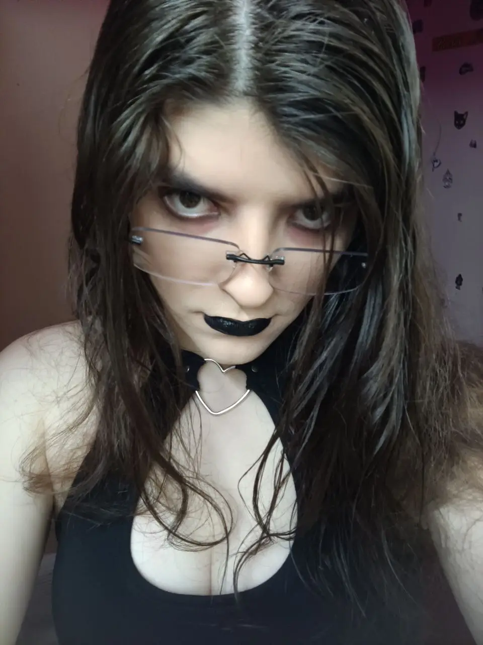 A person with long dark hair, heavy black eyeliner, and black lipstick stares intensely at the camera while wearing…