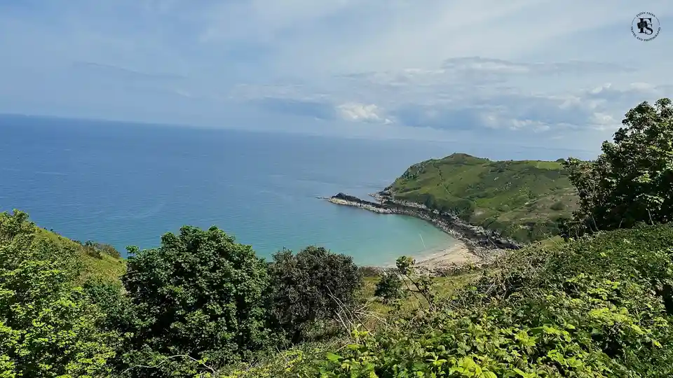 A scenic coastal view shows a bay with turquoise water nestled between green, rolling hills under a partly cloudy sky. The…