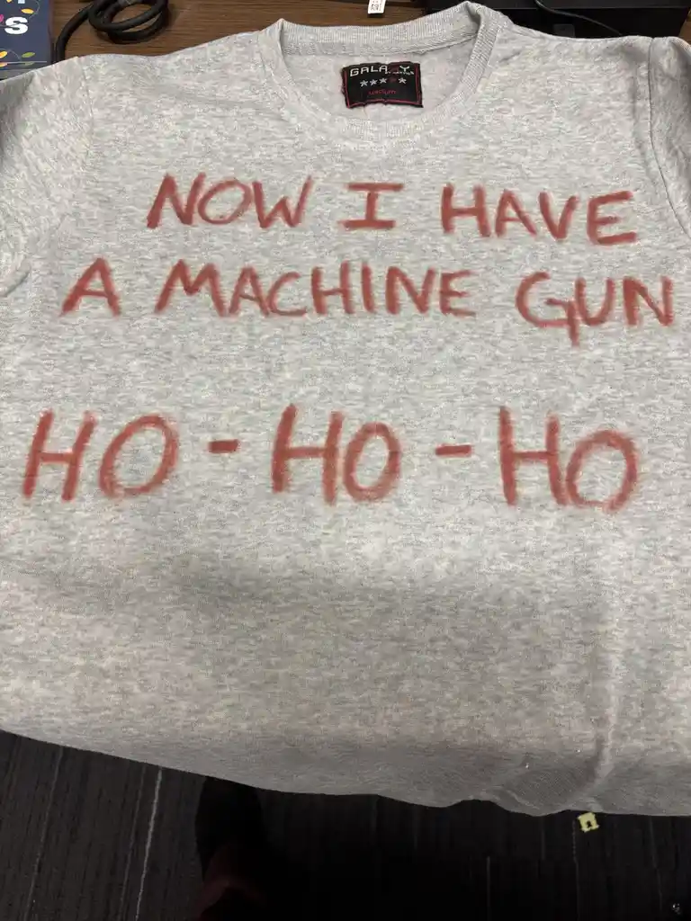 A gray sweatshirt laid flat has red handwritten text that reads 'NOW I HAVE A MACHINE GUN' on the top line and 'HO-HO-HO' on…